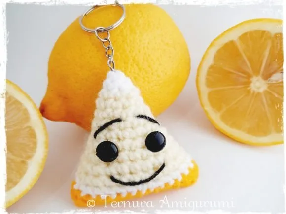 Crocheted lemon slice keychain with a smiling face, resting next to fresh whole and sliced lemons.